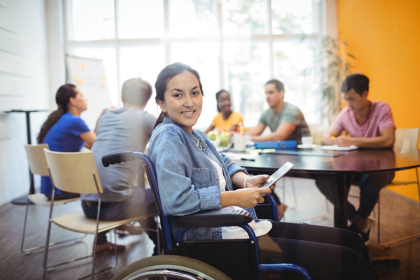 A young disabled woman in a wheelchair looks to the camera while others behind her sit at a table.