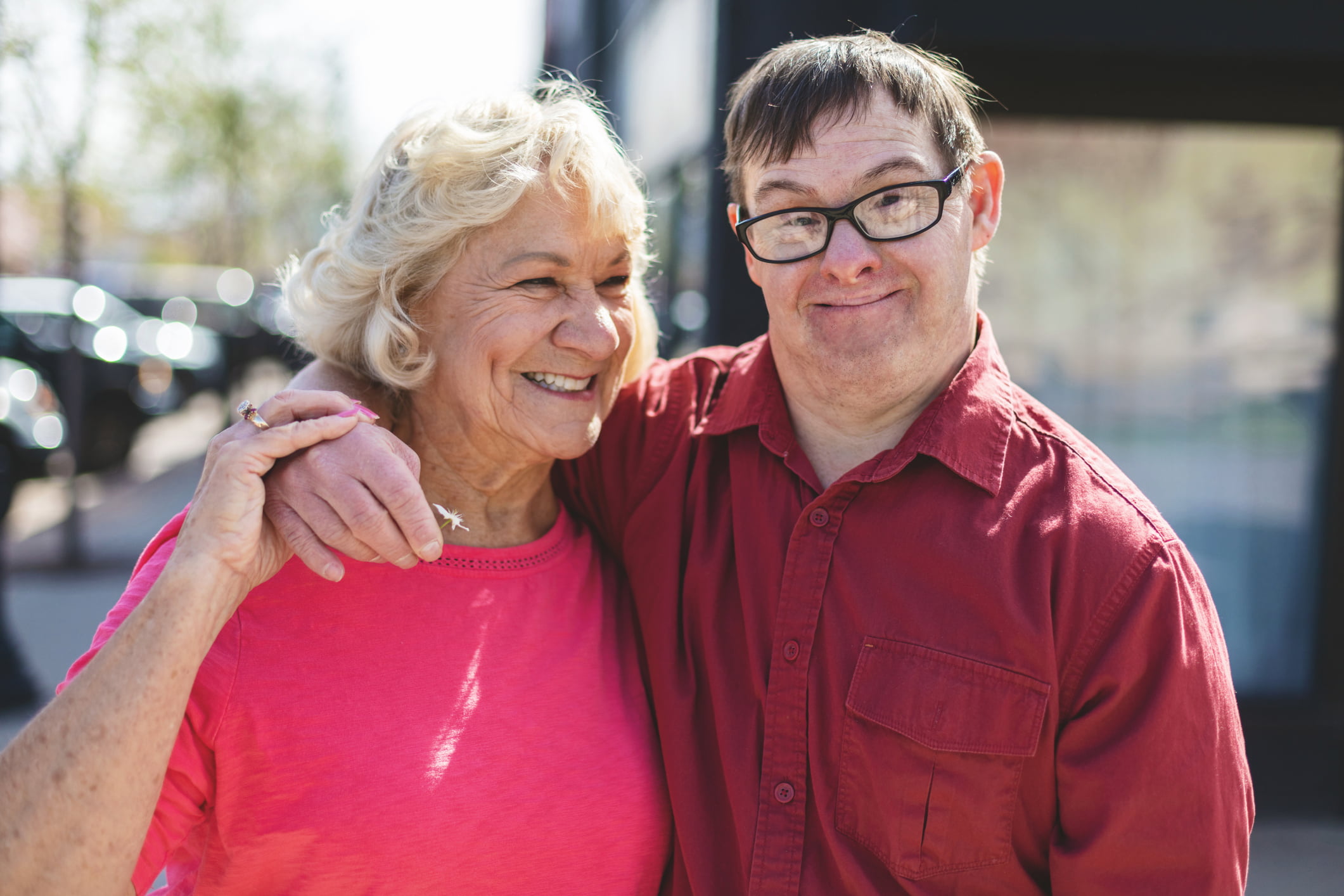A smiling Down Syndrome man stands with his arm around a smiling older woman.