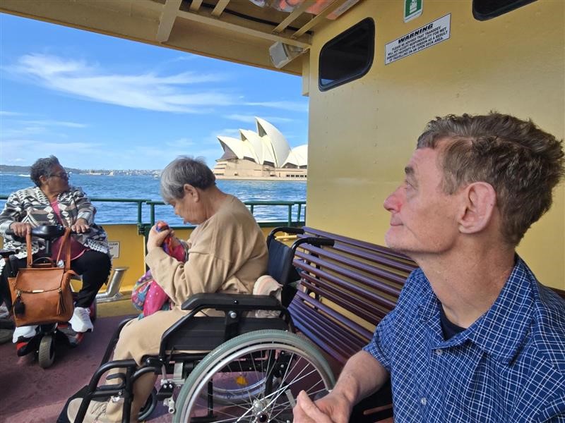 A group of people sit on a ferry, with the Sydney Opera House in view.